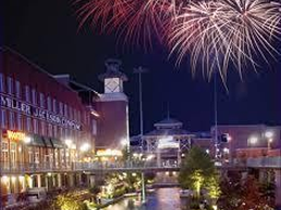Fireworks light up the night sky over a lively riverside area with buildings and a clock tower.