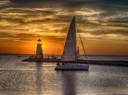 Sailboat glides past lighthouse at sunset.